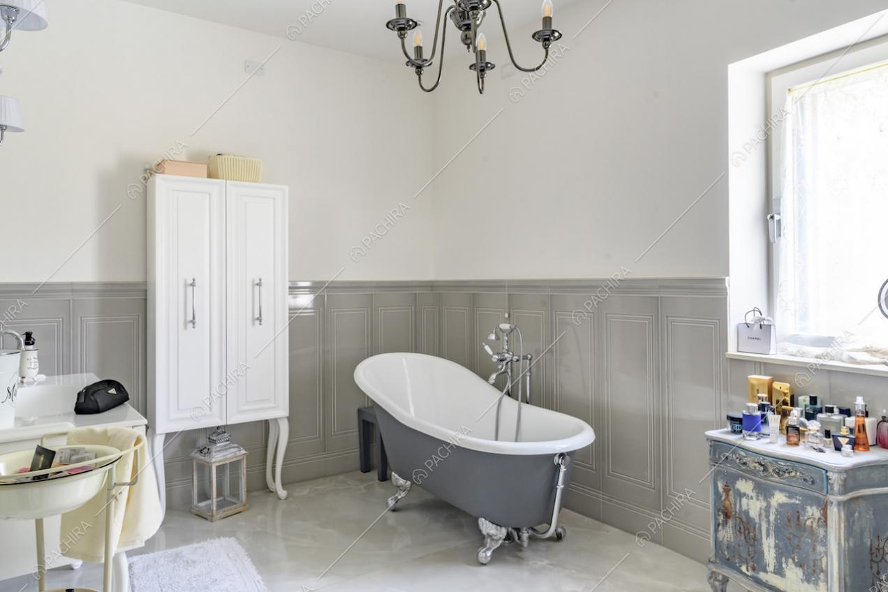 Bagno con vasca, pareti bianche e grigie con boiserie, pavimento in piastrelle e lampadario